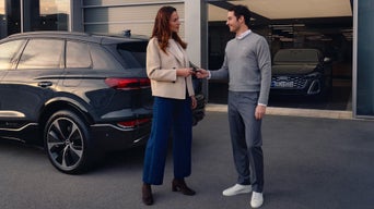 Mann übergibt Frau vor Autohaus den Autoschlüssel eines grauen Audi SUV auf einem Parkplatz.