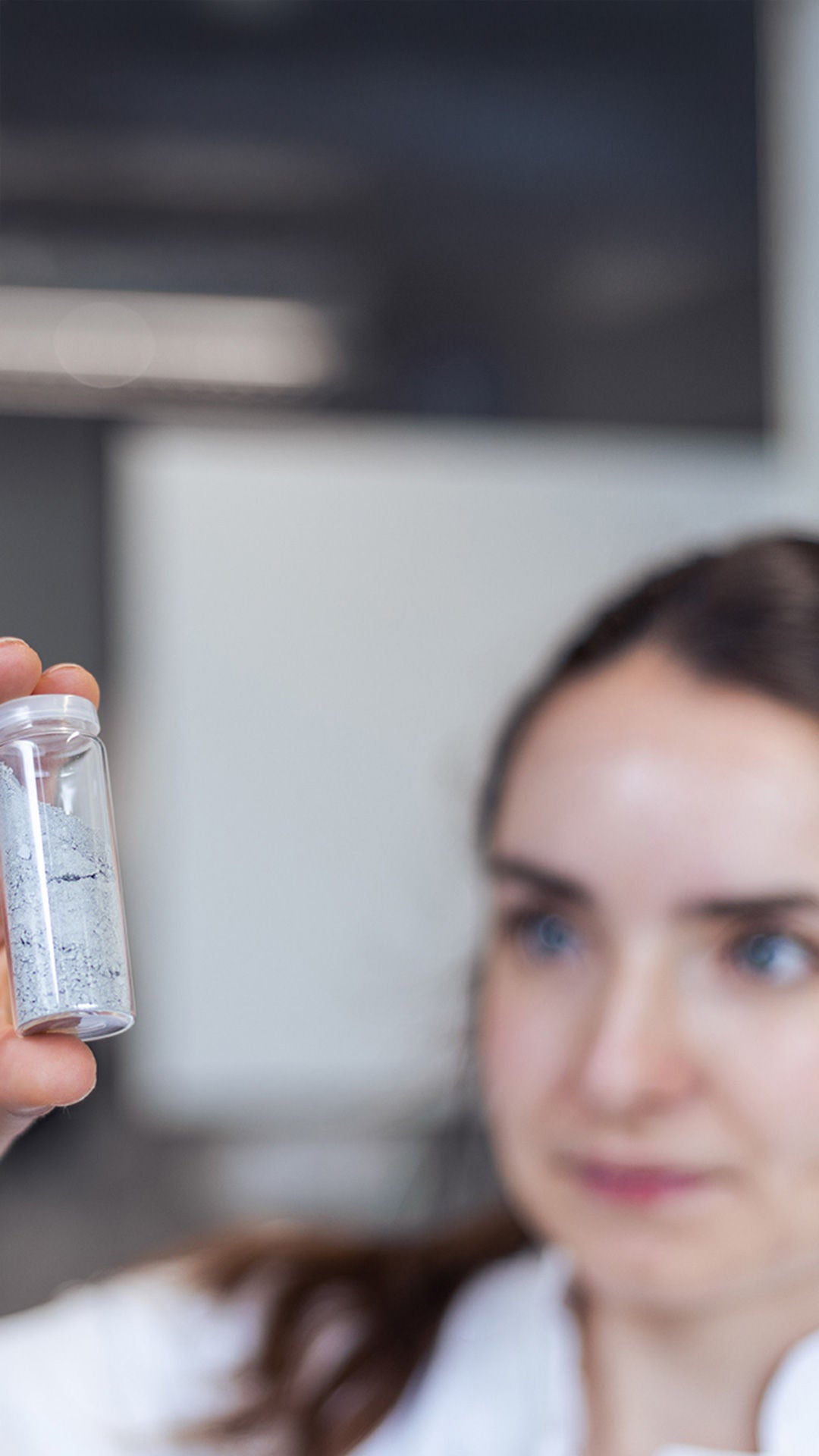 Frau in einem weissen Arbeitskittel blickt in ein Fläschen mit einer Füllung.