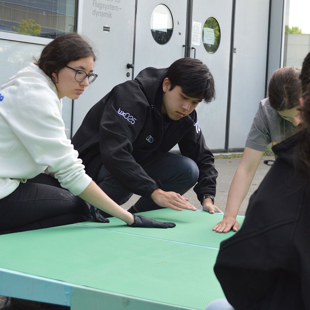 A few students work on a green cardboard lying on the floor