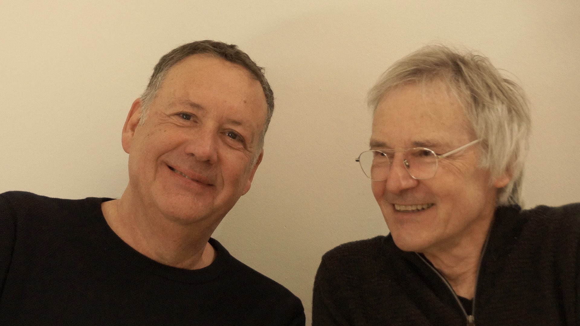 Thomas Faist und Heinz Frommeyer posieren in dunklen Oberteilen vor einer weißen Wand.