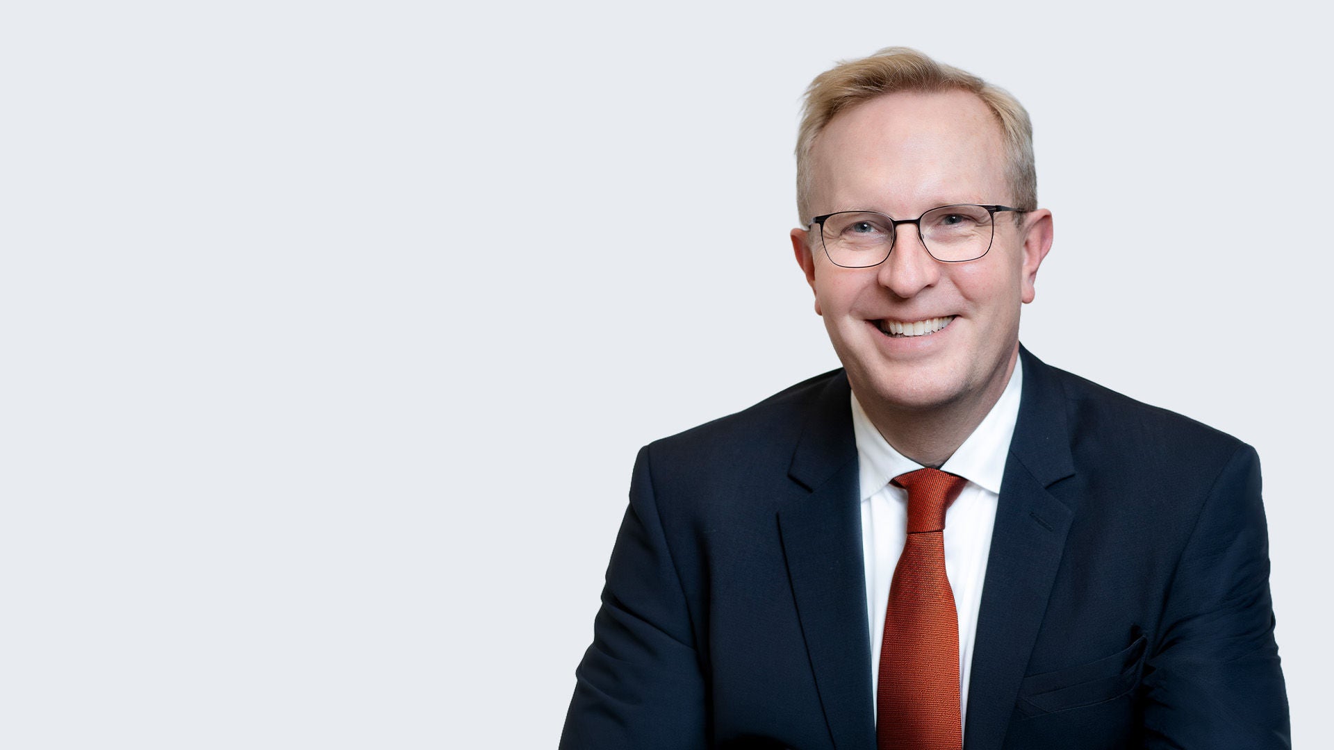 Lächelnder Mann mit Brille, dunklem Anzug und roter Krawatte vor hellgrauem Hintergrund.