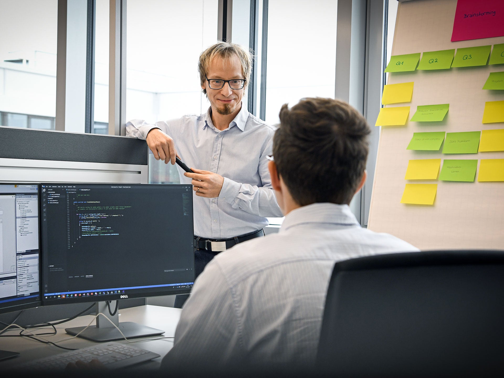 Two people discussing in an office. One stands near a whiteboard covered with sticky notes, the other sits at a desk with a computer monitor