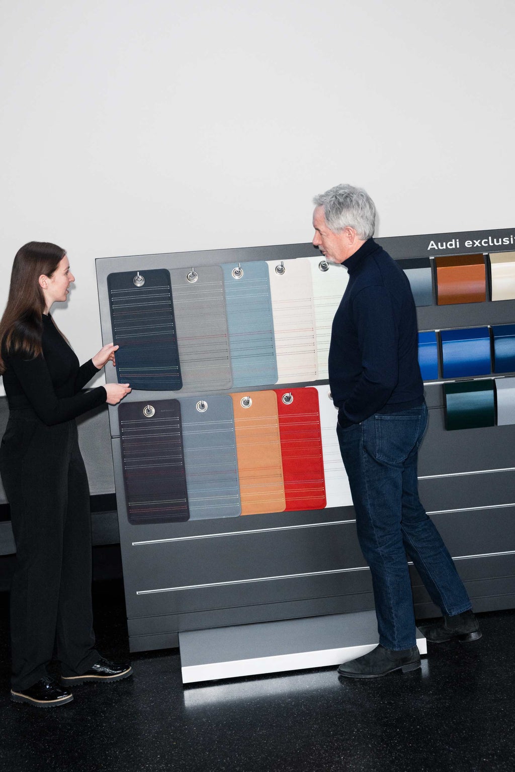 Annika Stößl und Trevor Hill unterhalten sich an der Audi exclusive Musterwand, an der Interieur und Exterieur Farbmuster zu sehen sind.