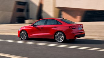 Rote Audi A3 Limousine fährt auf zweispuriger Straße vor moderner Gebäudefassade entlang