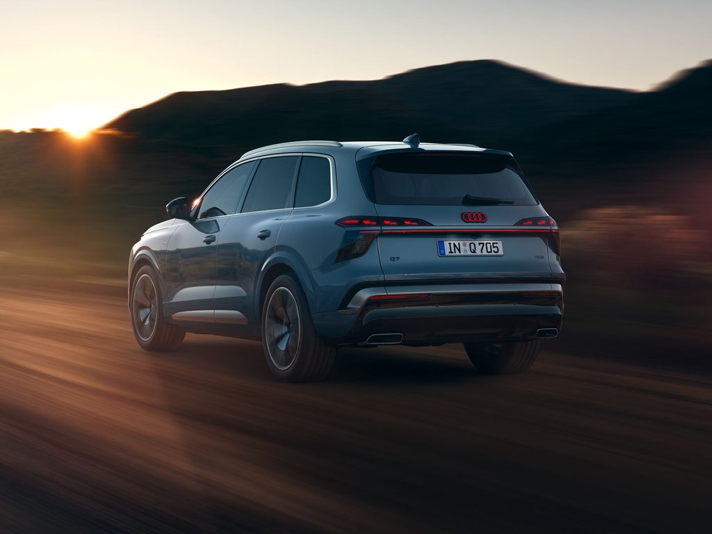 Le SUV Audi Q7, vu de biais depuis l'arrière alors qu'il roule à toute vitesse sur une route de campagne, avec les montagnes en arrière-plan. Le soleil couchant à l'horizon illumine le paysage.
