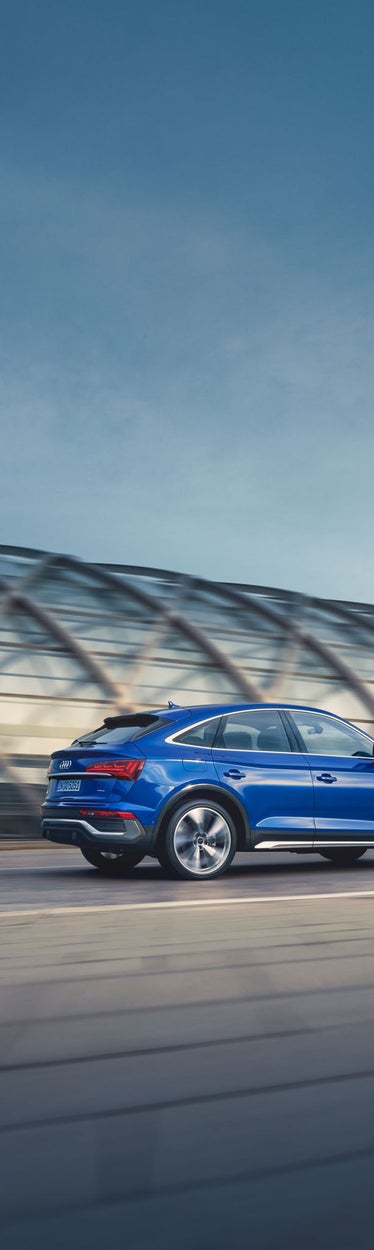 Blauer Audi Q5 Sportback fährt auf Straße, modernes Gebäude mit Glasstruktur im Hintergrund.