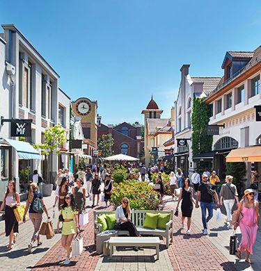Viele Menschen schlendern durch eine sonnige Outlet-Shoppingmeile mit bunten Geschäften und Pflanzen.