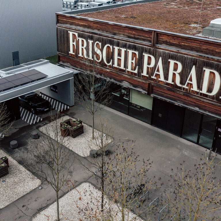 Ein Audi lädt an einer überdachten Ladesäule neben einem Gebäude mit großem Schriftzug „Frische Paradies“.