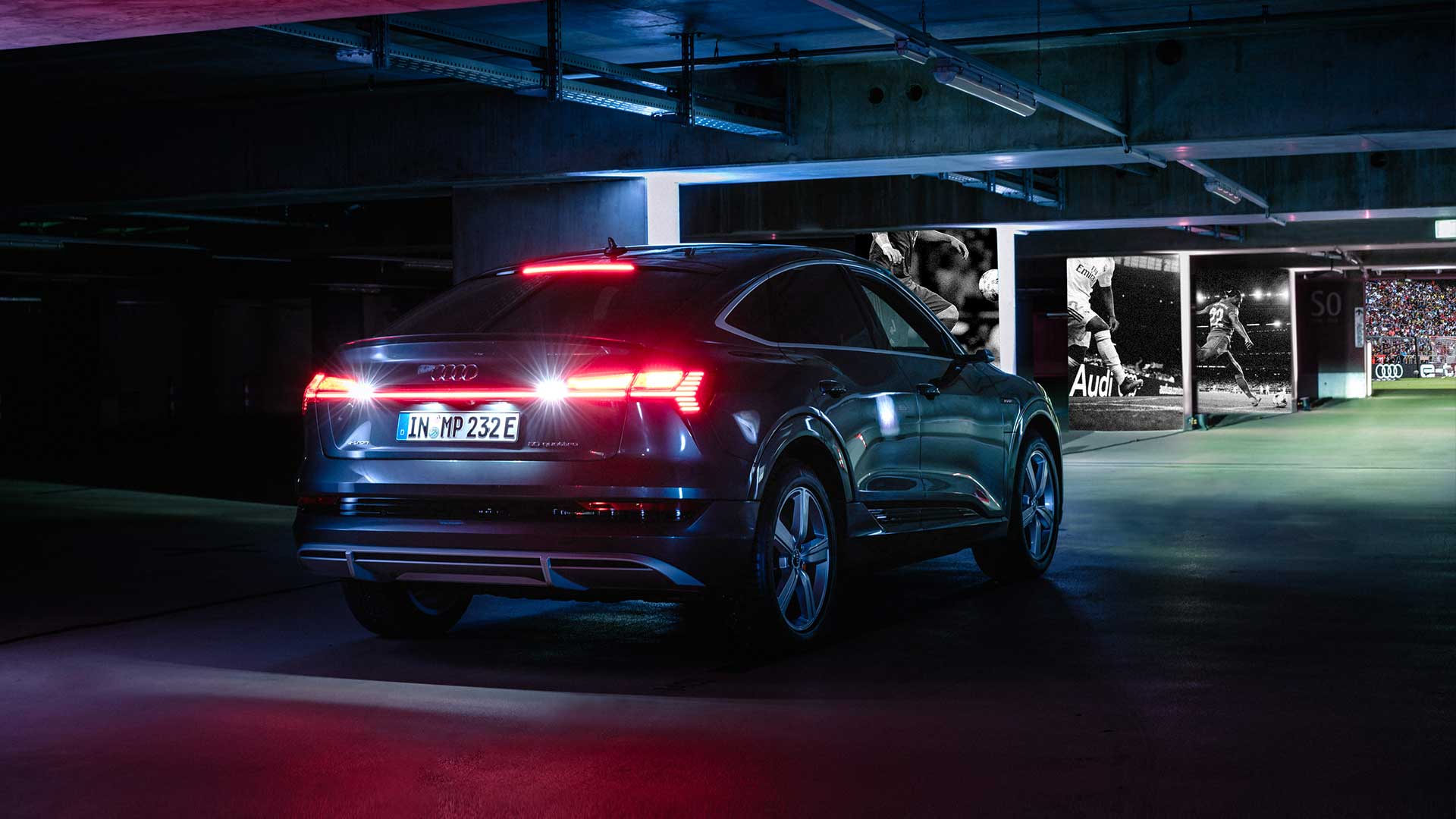The rear lights of an Audi driving through the underground car park with football motifs on the walls towards a door behind which the inside of a stadium with full stands can be seen.
