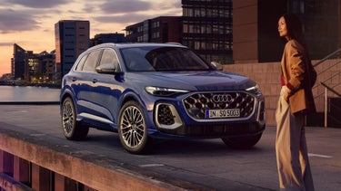 Der SQ5 SUV steht vor der Skyline einer Küstenstadt. Davor befindet sich eine junge, modische Frau.&nbsp;