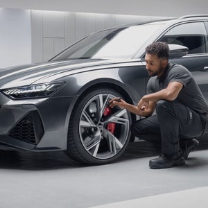 A mechanic inspects the wheel and brake system of a sleek grey Audi sports car in a modern, well-lit garage.