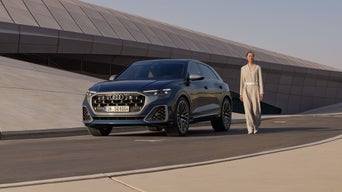 De SQ8 staat geparkeerd voor een futuristisch gebouw met een gebogen vorm, waarnaast een modieus geklede jonge vrouw staat.