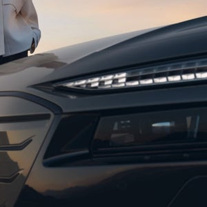 A person in a white jacket stands beside a sleek black car, showcasing its modern design against a soft sunset backdrop.