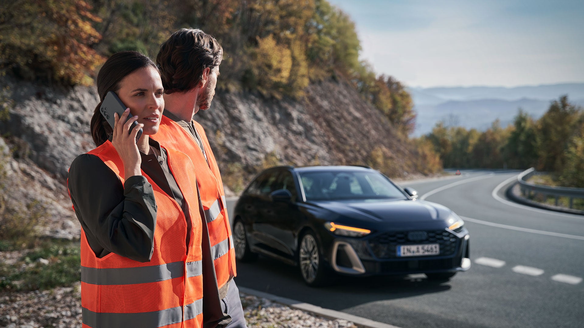 Ein Paar in Warnwesten steht am Straßenrand neben ihrem schwarzen Audi, der eine Panne hat. Die Frau telefoniert am Handy.