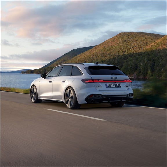 A sleek white Audi car drives along a scenic road by a lake with lush green hills in the background under a partly cloudy sky.