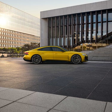 Gelber Audi RS e-tron GT performance geparkt vor einem modernen Gebäude mit Glasfassade und Treppe bei Sonnenuntergang.