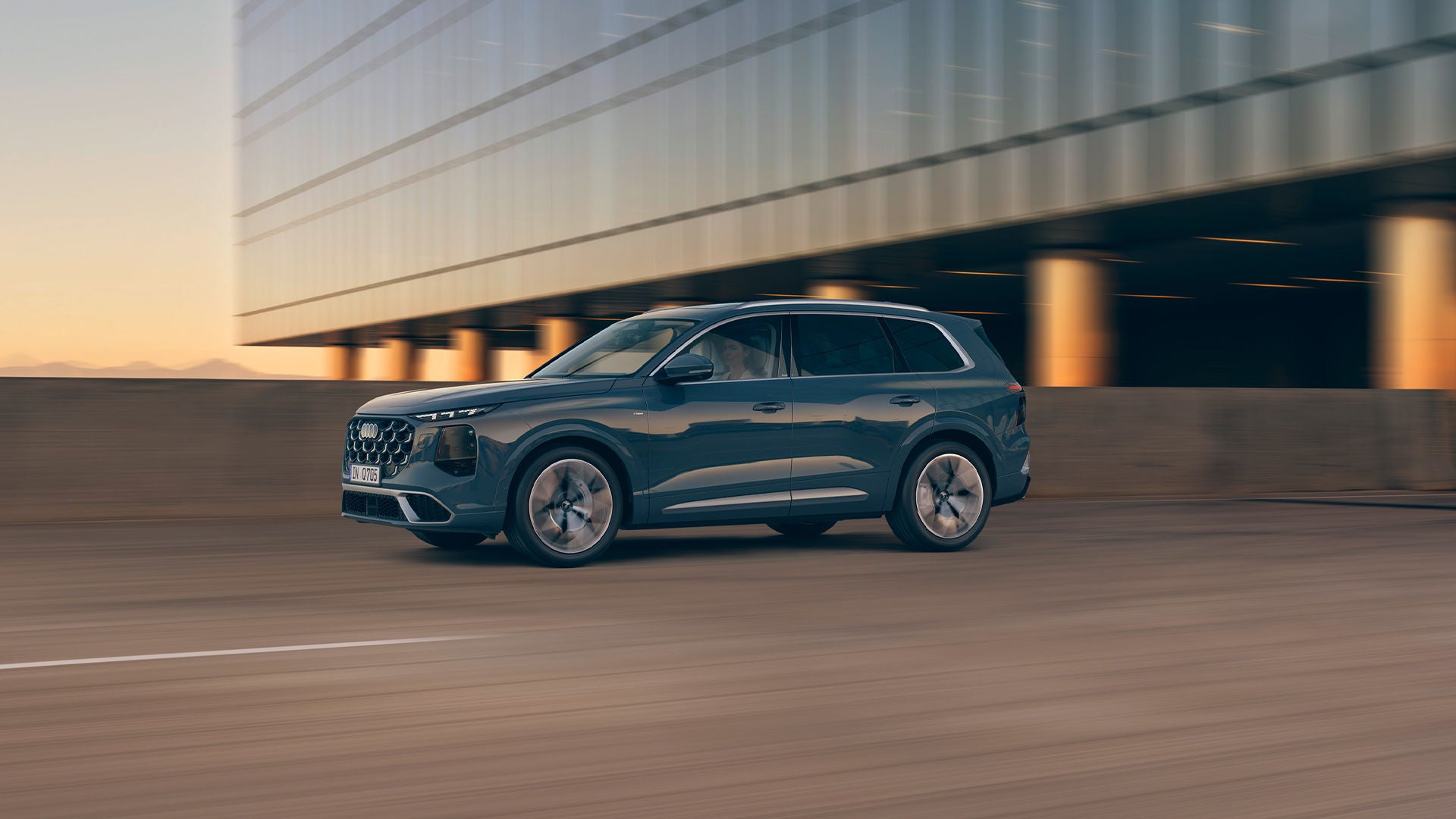 Une Audi Q7 grise roulant au coucher du soleil devant un immeuble moderne.