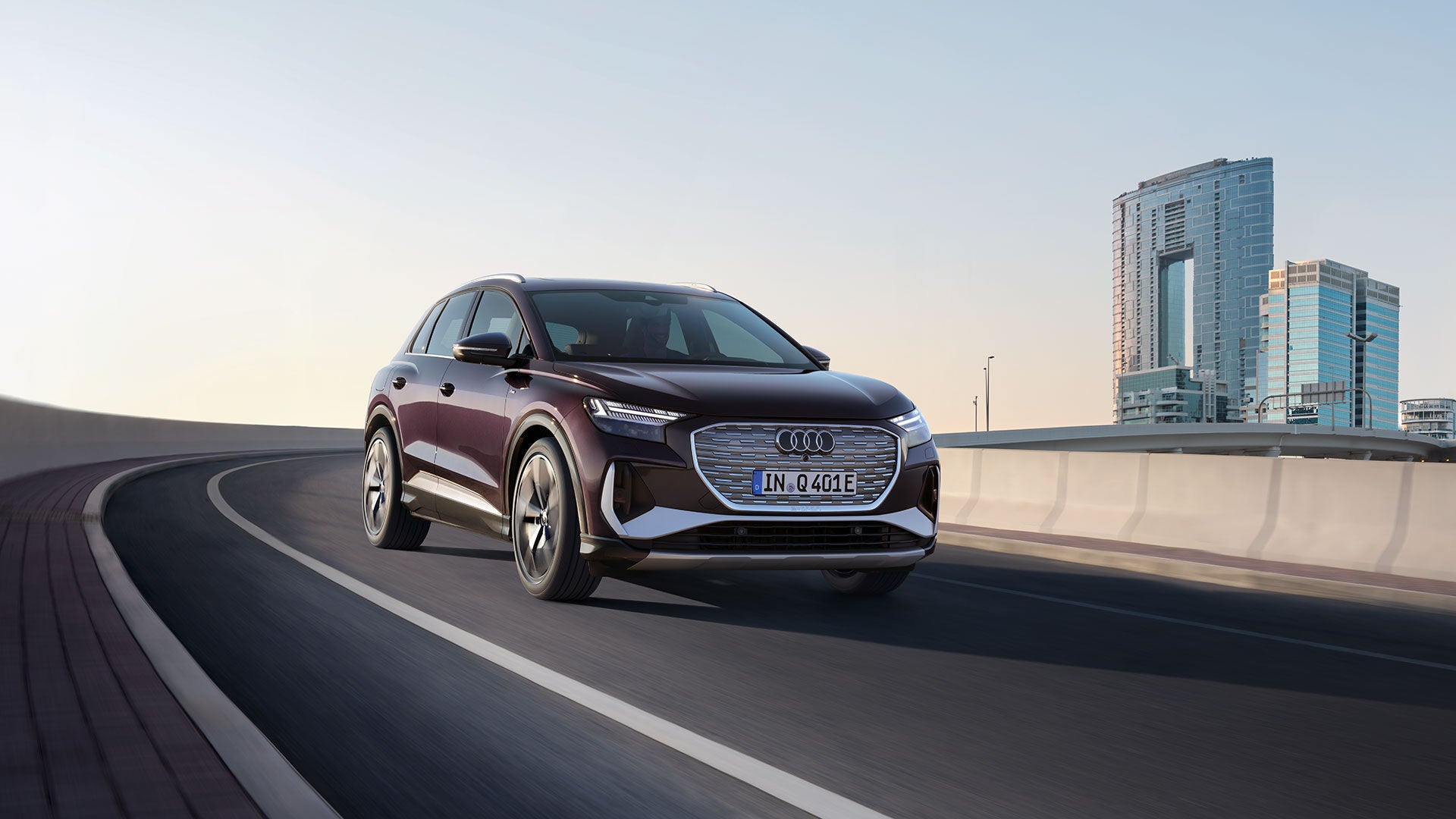 Ein dunkelroter Audi Q4 e-tron fährt auf einer leeren, kurvigen Straße vor modernen Hochhäusern bei klarem Himmel.