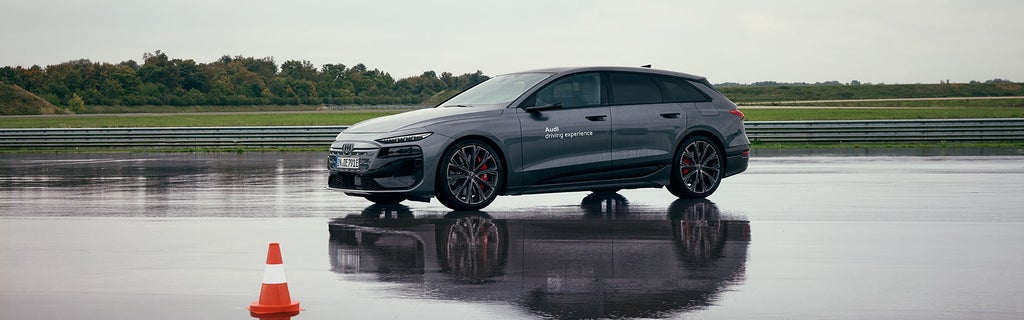 Ein grauer Audi S6 Avant e-tron mit der Aufschrift „Audi driving experience“ steht auf nasser Fahrbahn neben einem Verkehrshütchen.