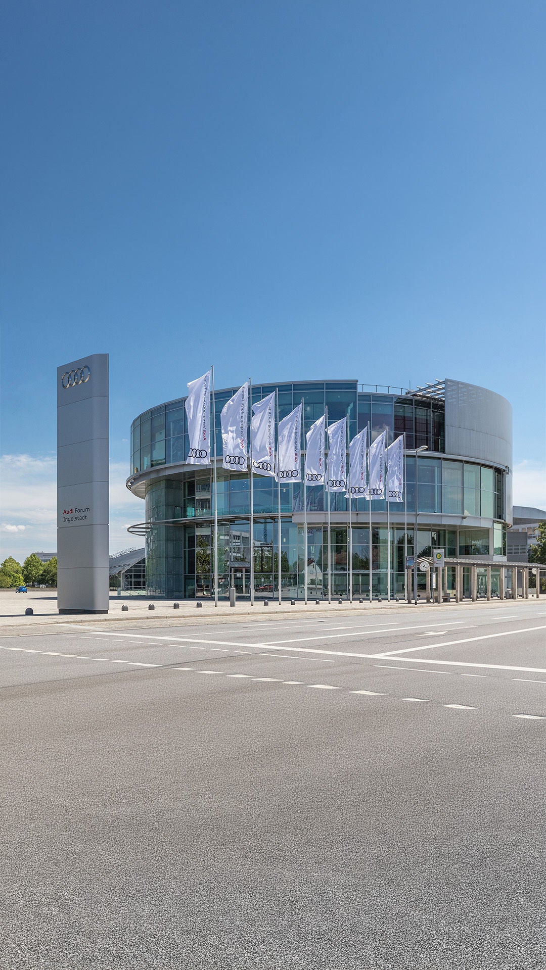 Modernes Glasgebäude des Audi Forum Ingolstadt mit Audi-Flaggen und großem Logoschild vor blauem Himmel.