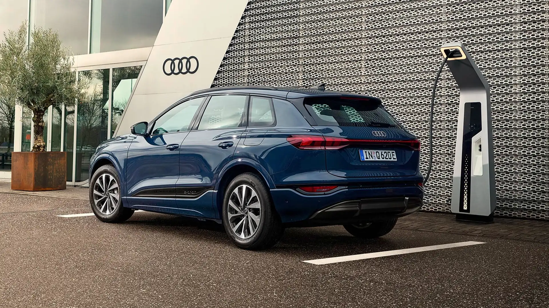 Blue Audi Q6 e-tron parked at a public charging station outside an Audi dealership, shown from the rear three-quarter angle. The SUV features a sculpted body design, large alloy wheels and a full-width rear LED light bar. A charging cable is connected to the vehicle, while the Audi rings logo, floor-to-ceiling windows and modern architectural details form the background.