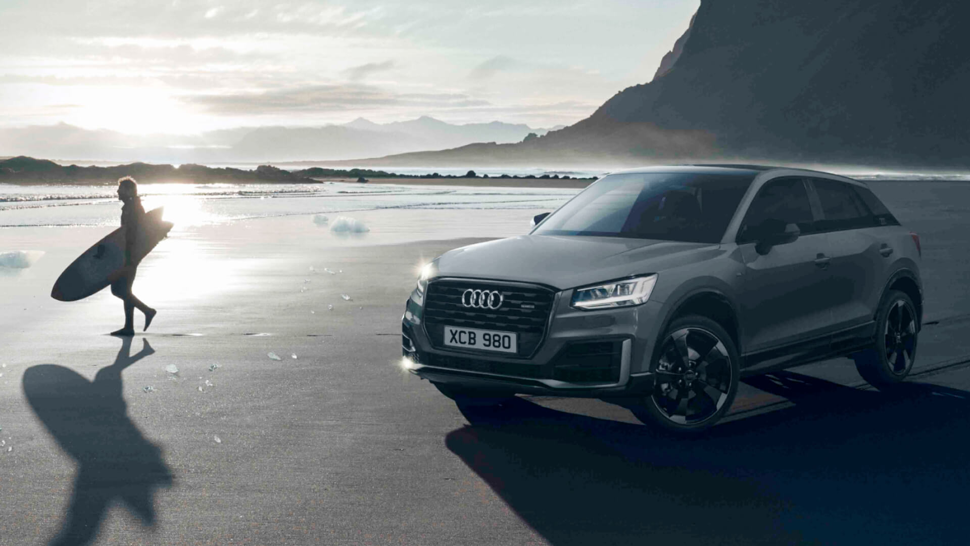 A beach in the late afternoon with a surfer walking on the left & a parked Audi vehicle on the right