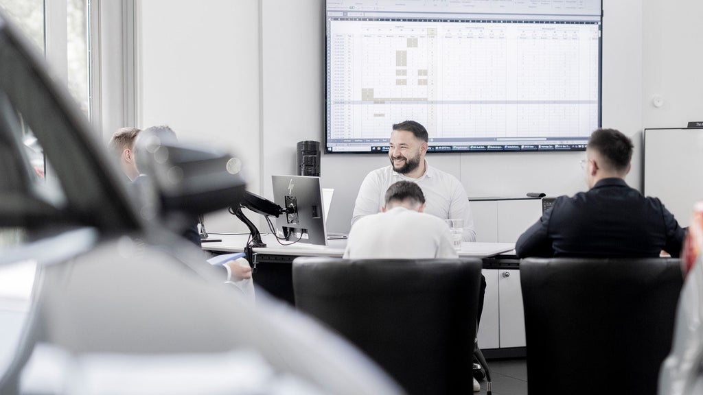 People are sitting at a table and talking. A screen with a document on it hangs on the wall.