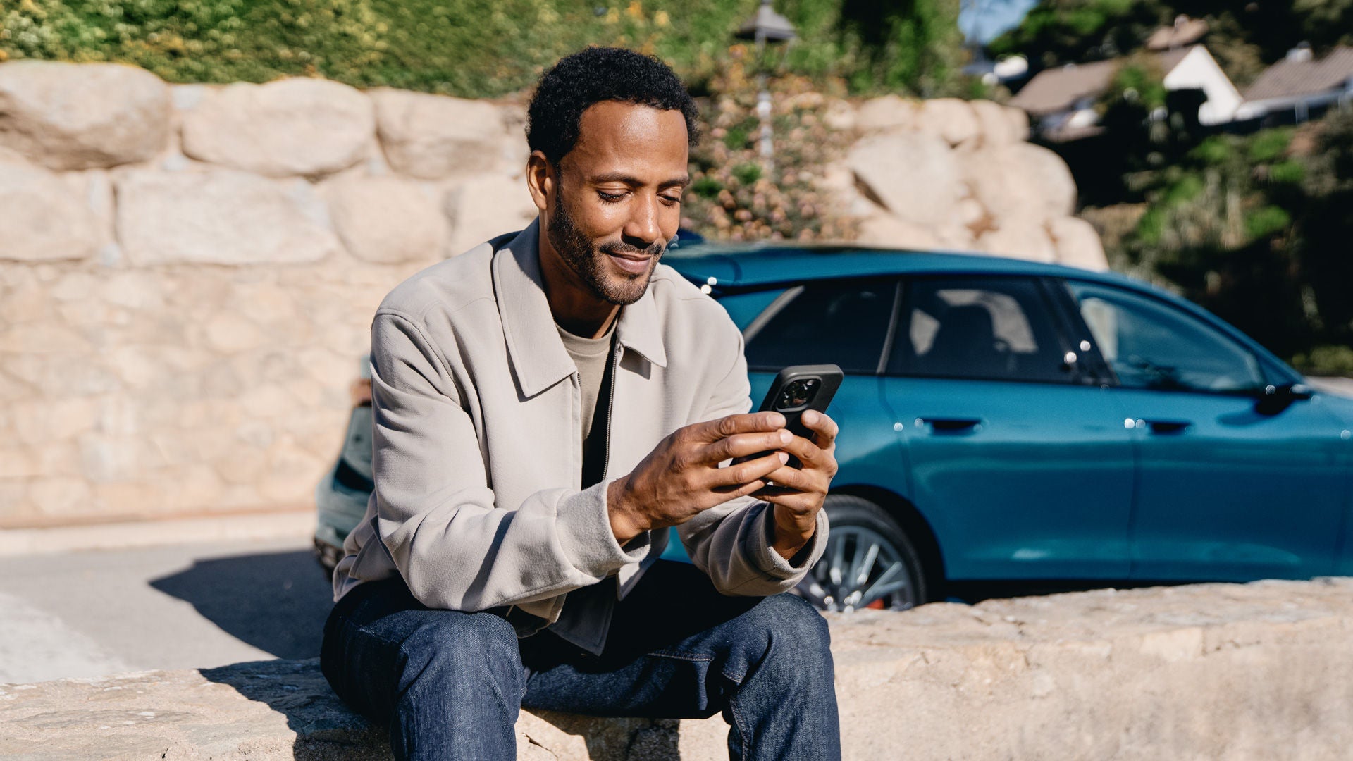Person sitzt im Freien auf einer Steinmauer und hält ein Smartphone in den Händen. Im Hintergrund steht ein blauer Audi.