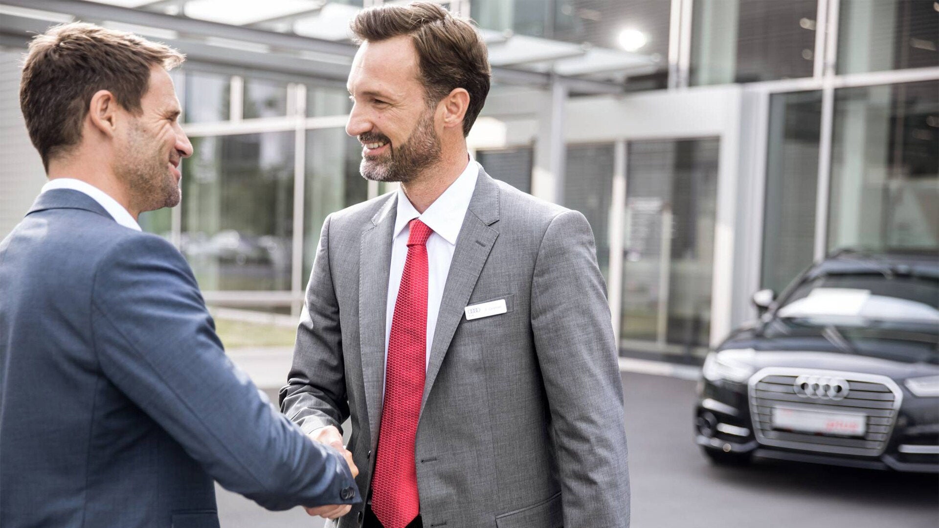 Zwei Männer im Anzug geben sich vor einem Audi-Gebäude die Hand und lächeln einander freundlich an.
