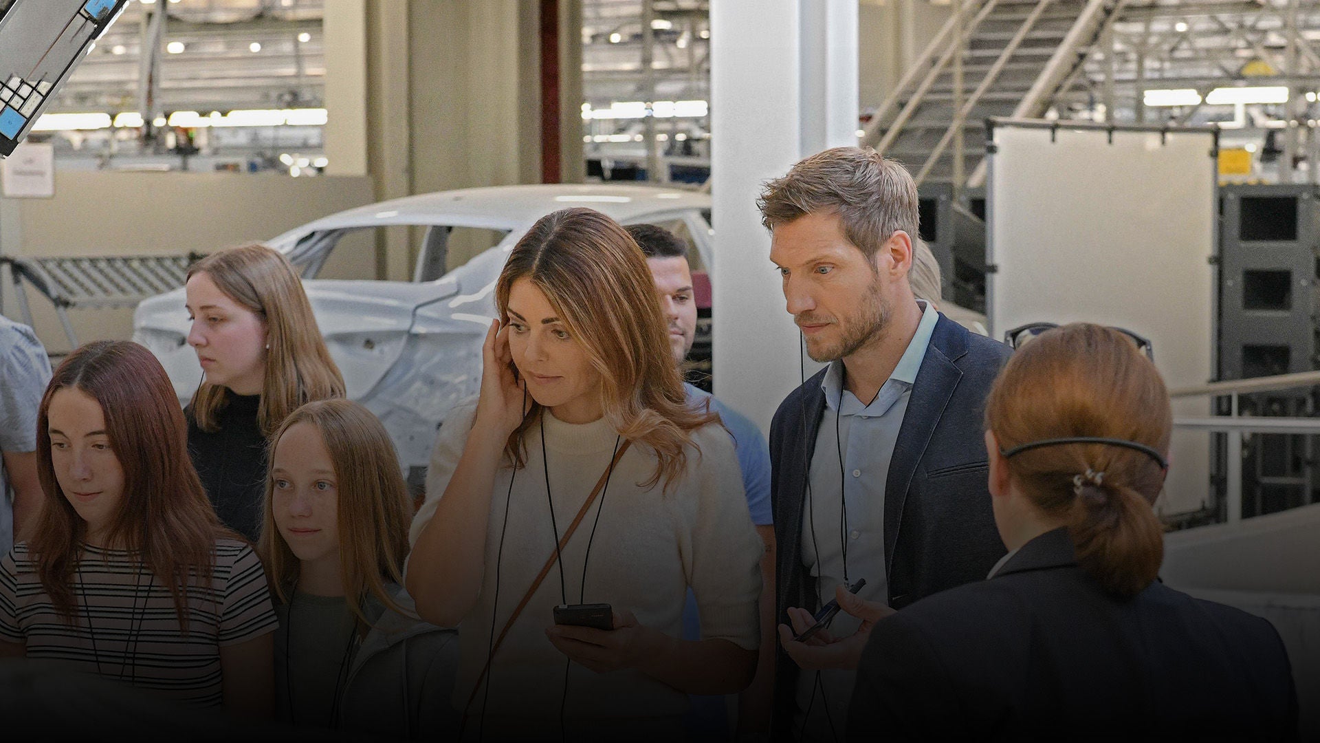 Group of people on a factory tour, man and woman in the foreground with headsets in the production hall.