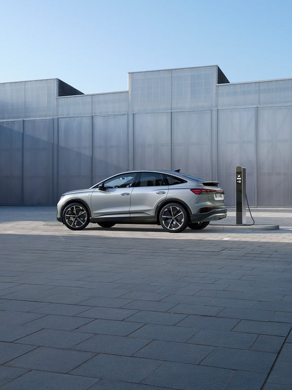 Silberner Audi Q4 Sportback e-tron lädt an Ladesäule vor grauer Gebäudefassade bei Tageslicht.