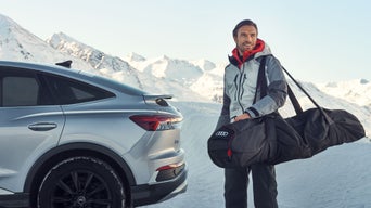 Mann in Winterkleidung trägt Audi-Skitasche neben silbernem Audi vor verschneiter Bergkulisse.