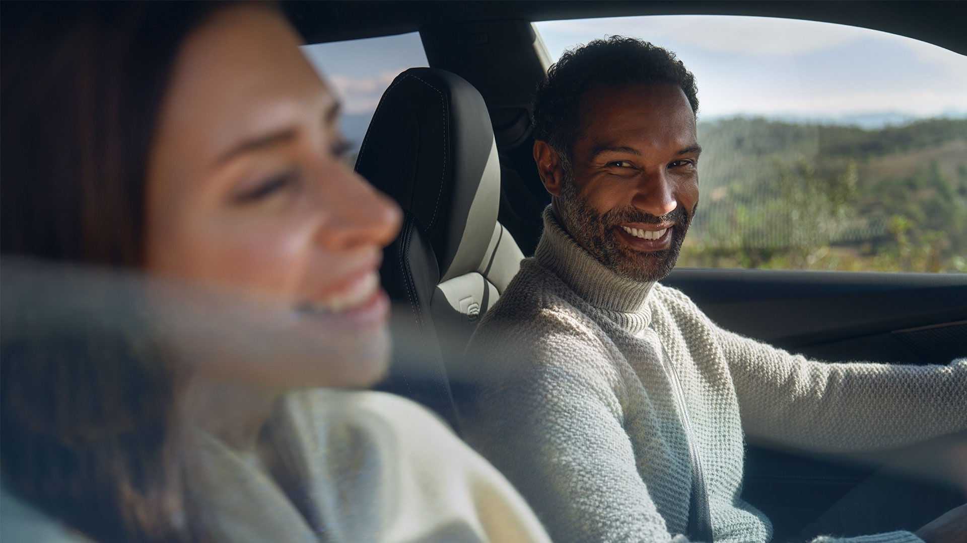 Deux personnes sont assises dans une voiture. La personne au volant regarde celle qui est assise à côté d'elle, que l'on aperçoit au premier plan, légèrement floue. À travers les vitres latérales, on distingue un paysage champêtre baigné par la lumière du jour.