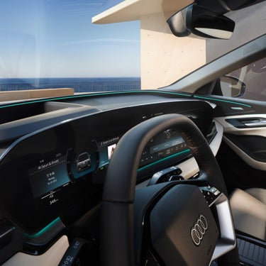 Digitales Cockpit eines Audi Q6 SUV e-tron mit Lenkrad, Touchscreen-Anzeigen und Meerblick durch die Frontscheibe.