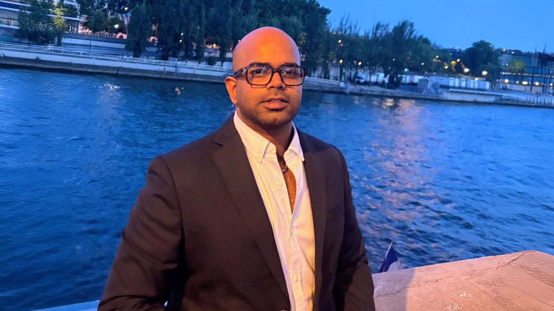 Mann mit Brille im schwarzen Anzug und weißem Hemd, neutraler Ausdruck, vor einem Gewässer.