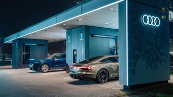 Zwei Elektroautos laden bei Nacht an modernen Audi-Ladestationen unter einem überdachten Ladehub mit Audi-Logo.