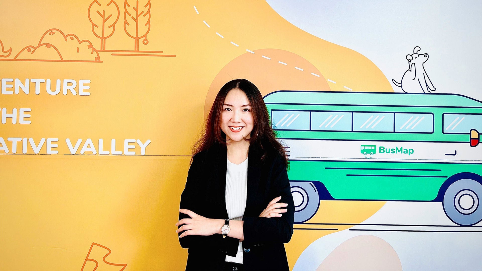 Frau mit langen dunklen Haaren im schwarzen Blazer, lächelnd, vor bunter Wandgrafik mit Bus und Landschaft.