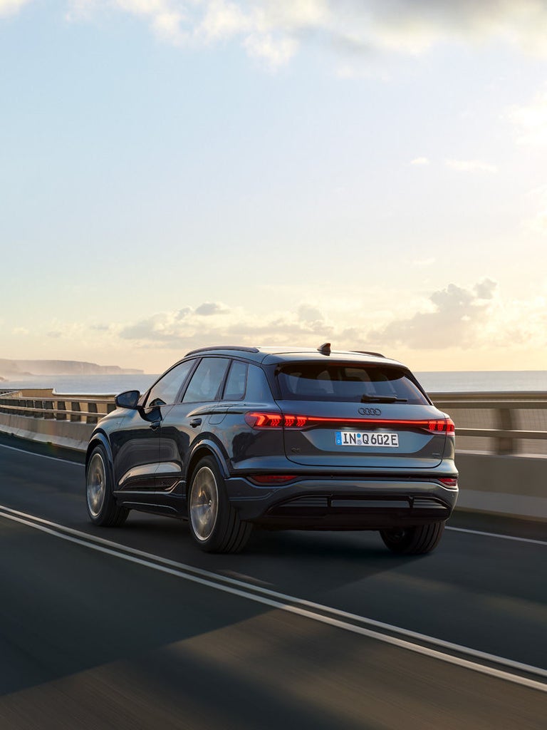 Ein blauer Audi Q6 SUV e-tron fährt bei Sonnenuntergang auf einer kurvigen Straße entlang der Küste.