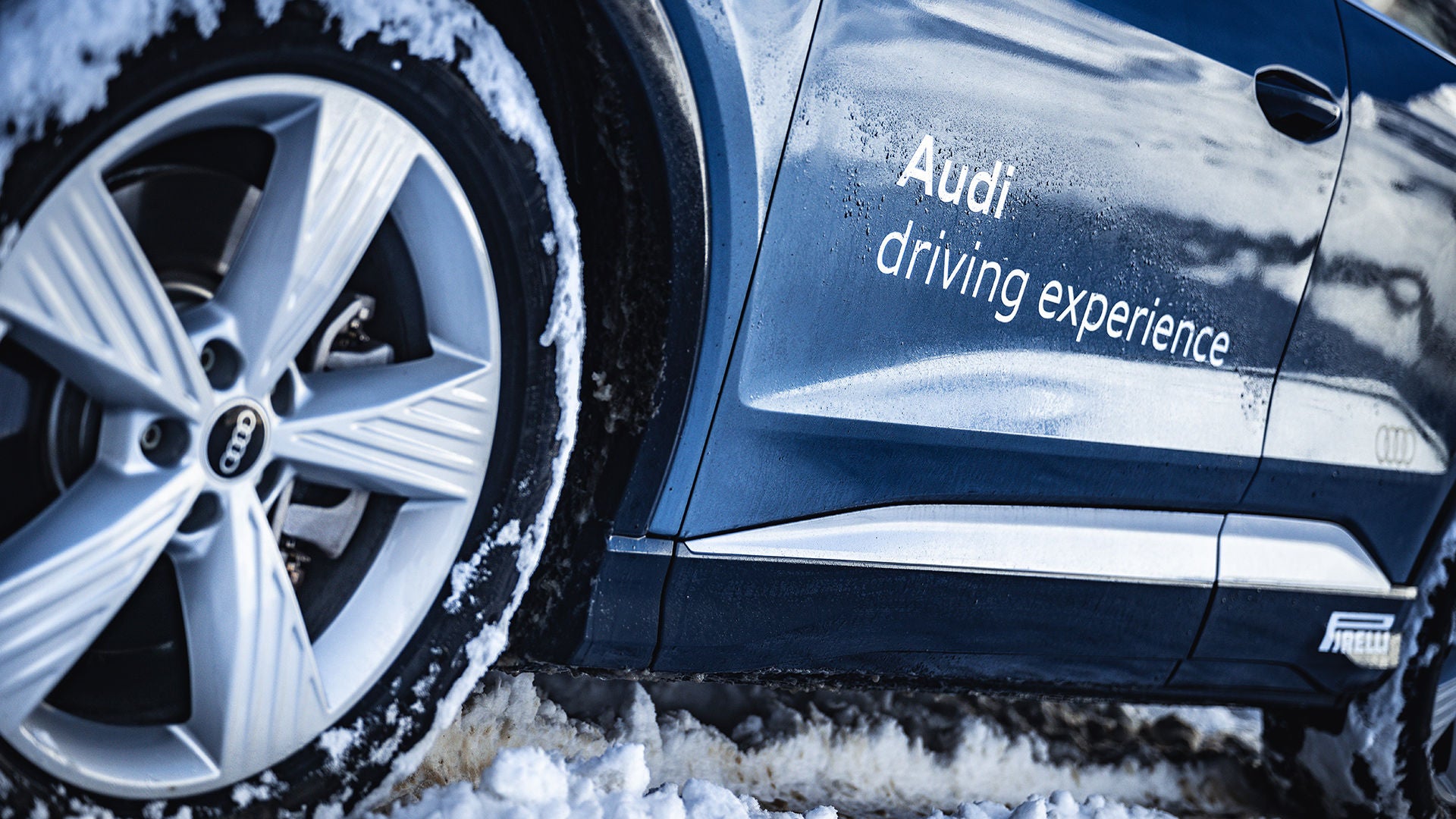 Dunkelblaues Audi Modell mit weißer Audi driving experience Aufschrift und Winterreifen von Pirelli im Schnee (Ansicht nur von Pirelli Reifen und Modellseite unten).