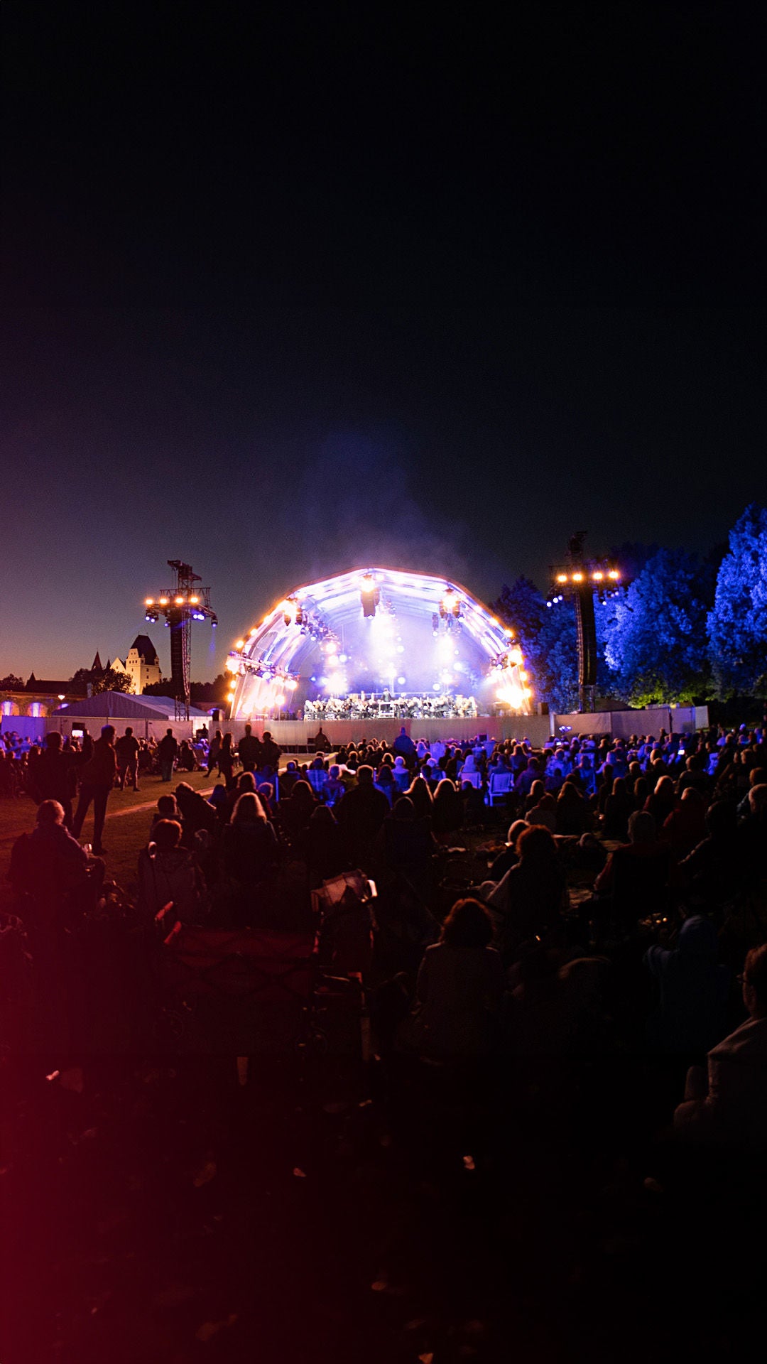 Im Vordergrund ist schemenhaft das Publikum der Audi Sommerkonzerte in der Dunkelheit zu erkennen. In der ferne befindet sich hell erleuchtet die Konzertbühne der Audi Sommerkonzerte.
