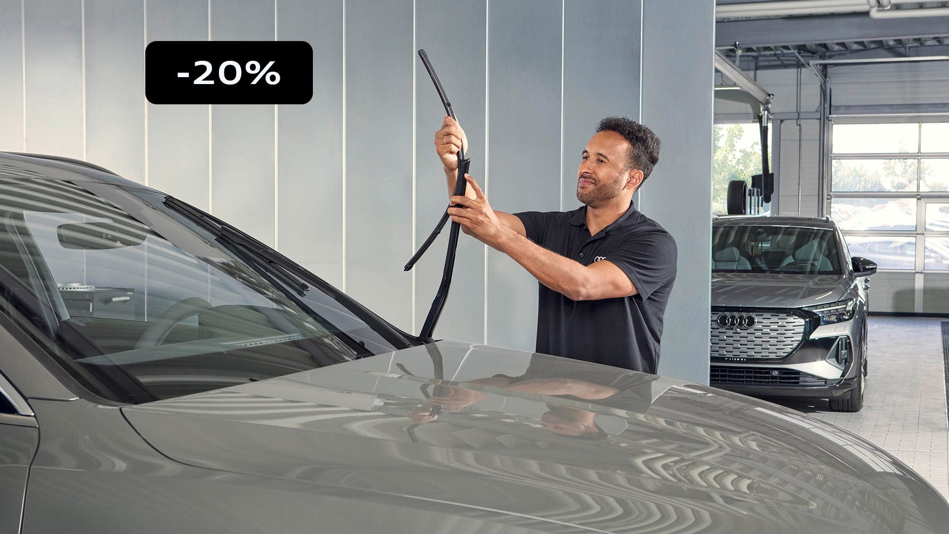 Personne dans un atelier automobile en train de relever un essuie-glace sur le pare-brise d’une voiture grise. Un encart noir indiquant « -20 % » apparaît dans le coin supérieur gauche, tandis qu’un autre véhicule est visible en arrière-plan.