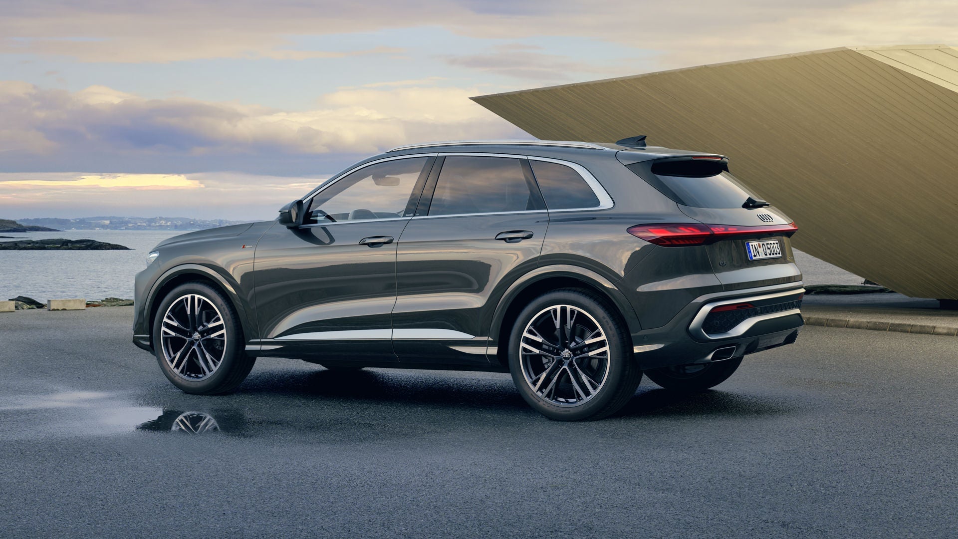 Der Q5 SUV steht im Seitenprofil auf einem Parkplatz direkt an der Küste. Daneben ein futuristisches Haus.