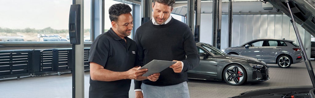 Zwei Männer stehen in einer Autowerkstatt neben einem geöffneten Auto mit sichtbaren orangefarbenen Hochvoltkabeln. Der linke Mann trägt ein schwarzes Polohemd mit Audi-Logo und hält ein Tablet, auf das beide schauen. Im Hintergrund sind zwei silberne Audi-Fahrzeuge zu sehen.