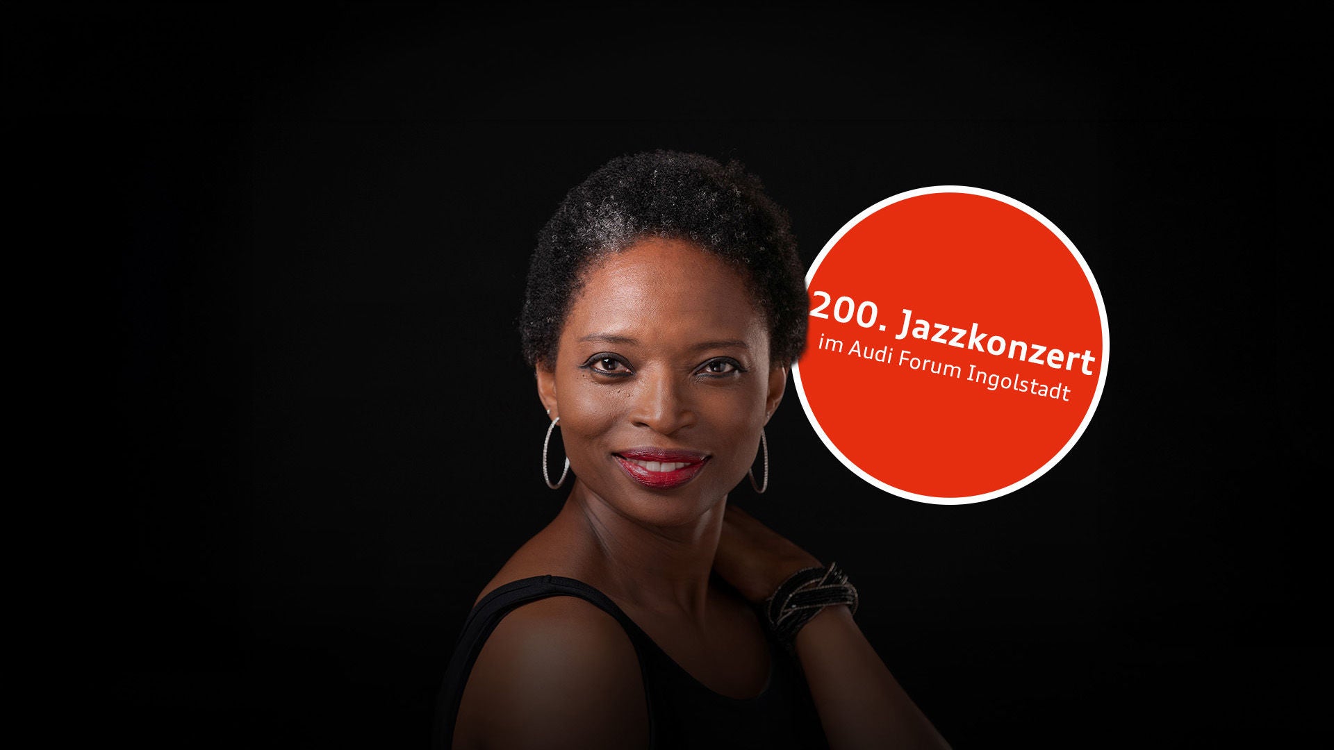 Porträt einer lächelnden Frau mit kurzem Haar, roten Lippen und Creolen; schwarzes Oberteil vor dunklem Hintergrund.