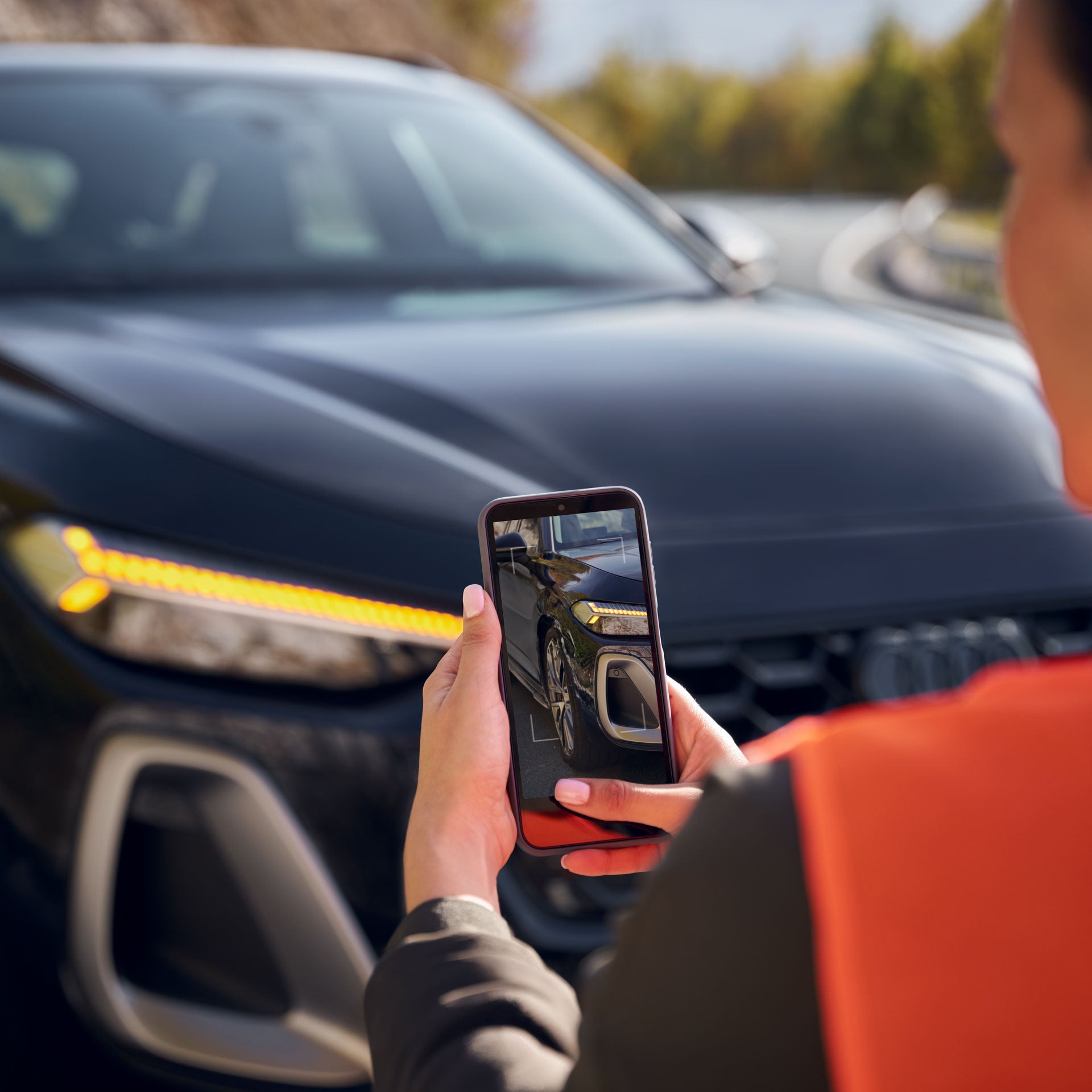 Person hält ein Smartphone und fotografiert die Front eines dunklen Autos auf einer Straße, LED-Scheinwerfer leuchten.