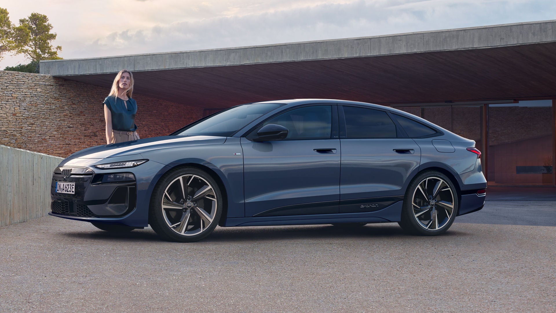 Der A6 Sportback e-tron steht im Seitenprofil vor dem Carport eines Hauses. Daneben steht eine junge Frau.