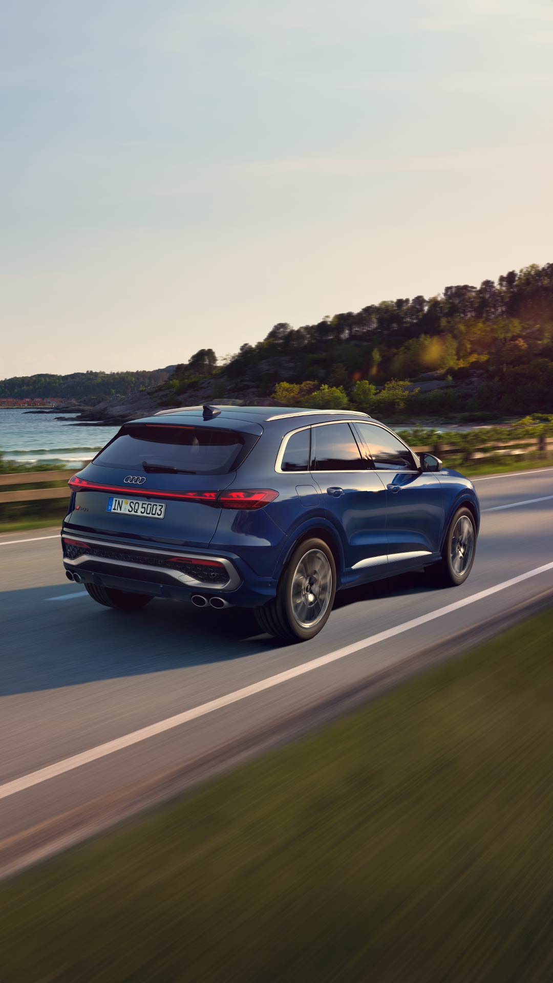 Ein blauer Audi SQ5 SUV fährt auf einer Küstenstraße bei sonnigem Wetter, der Schatten des Fahrzeuges ist sichtbar.