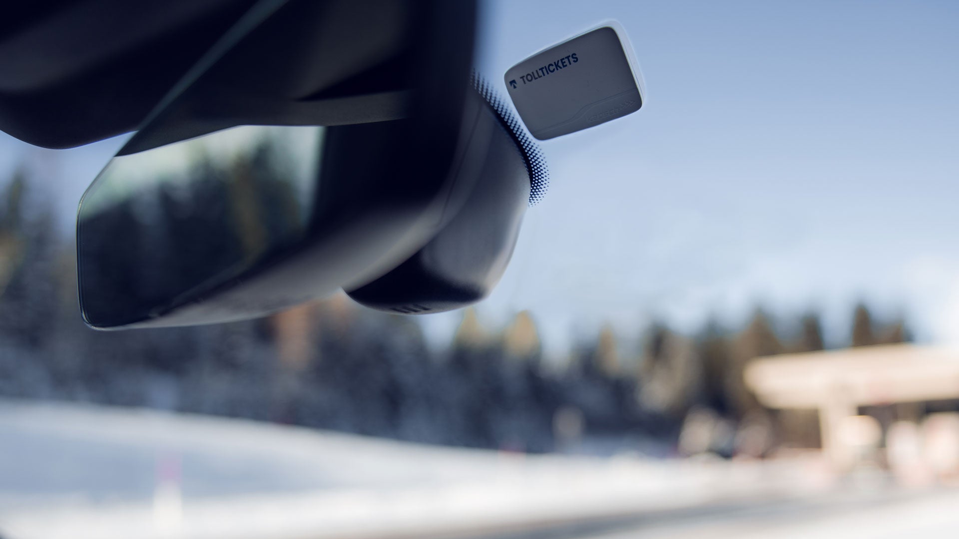 Kleine , weiße Mautbox klebt rechts hinter dem Rückspiegel an der Windschutzscheibe. Im Hintergrund ist verschwommen die Mautstation zu sehen.