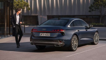 Blick in den Innenraum eines Audi mit digitalem Cockpit, farbige Frau in weißem Outfit geht außen an einer Betonwand vorbei.