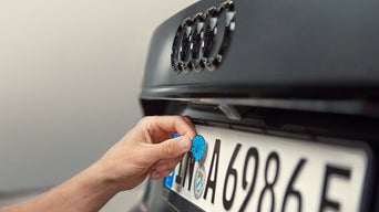 Eine Hand bringt eine blaue Plakette auf einem deutschen Nummernschild an einem schwarzen Audi an.